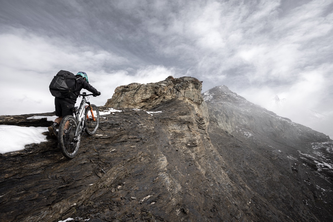 Matthew Fairbrother climbs a rocky ridge towards the summit with his bike. Snow covers parts of the path, while Ergon components (saddle and grips) provide stability.