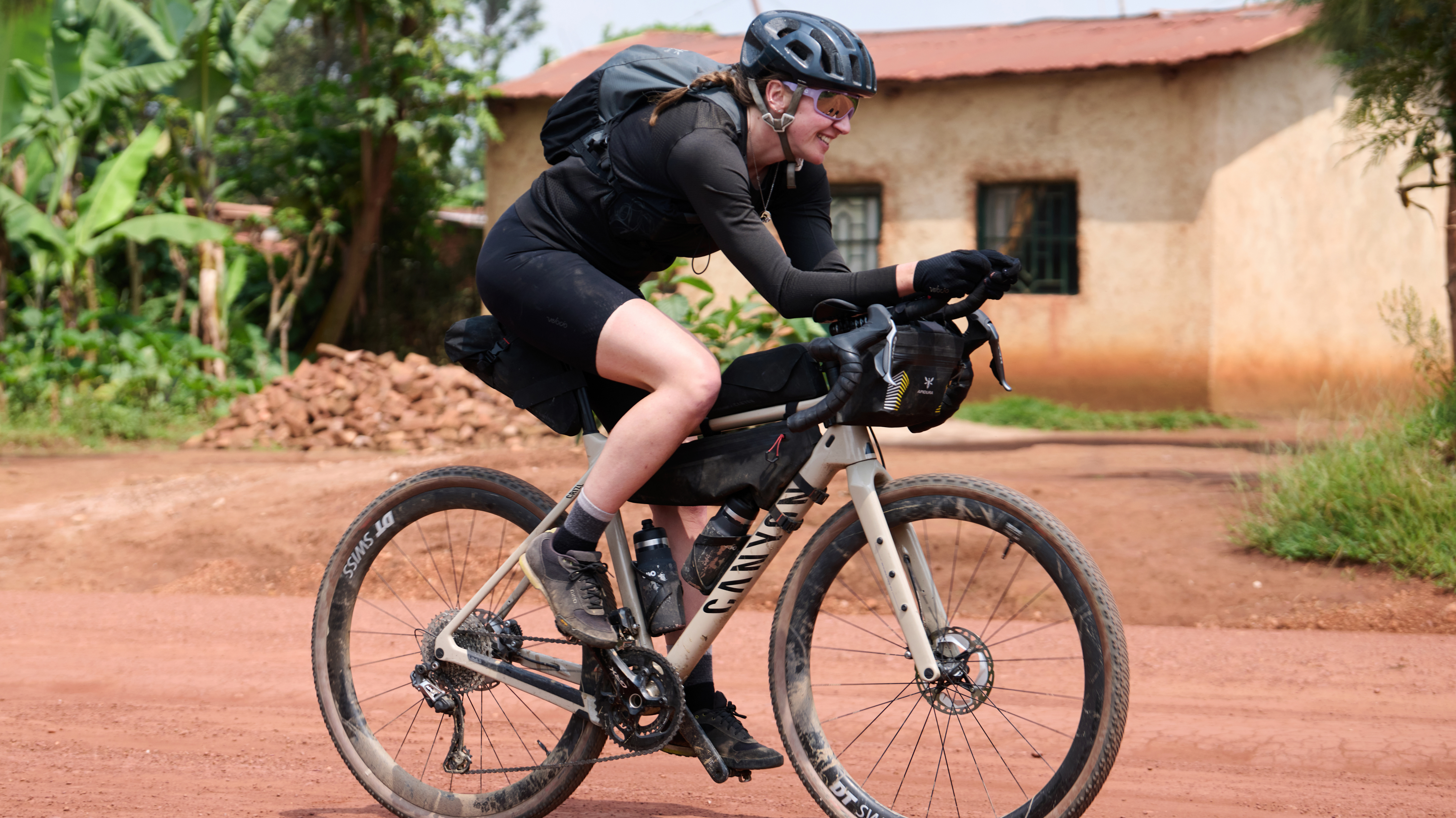 Jenny Tough fährt mit ihrem ihrem Gravelbike über eine unbefestigte Straße in tropischer Umgebung. Im Hintergrund stehen Palmen und ein Bungalow aus Lehm.