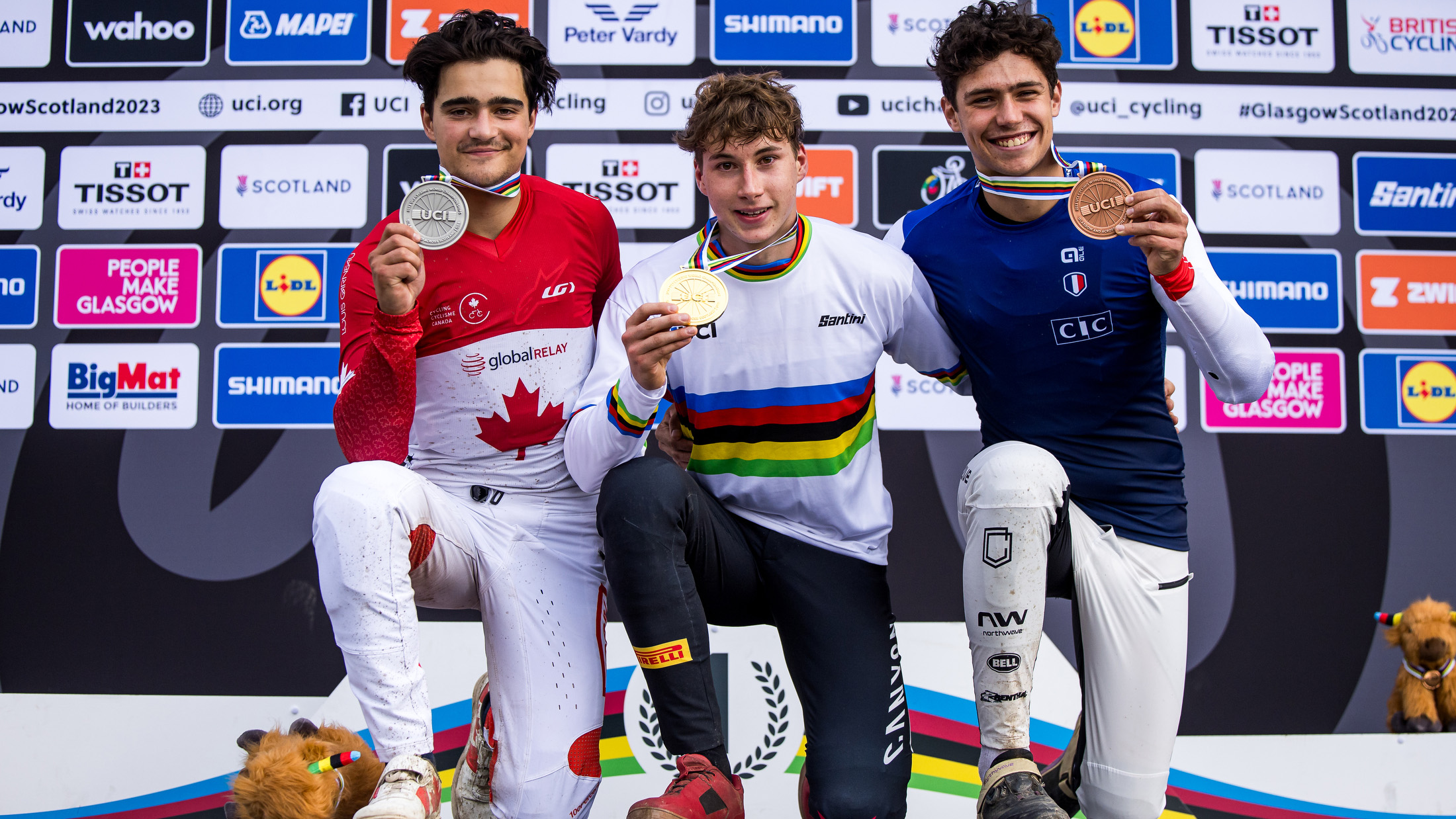 Henri Kiefer (Mitte) auf dem Podium nach seinem Sieg bei der UCI-Weltmeisterschaft. Er trägt das Regenbogentrikot des Weltmeisters und hält stolz seine Goldmedaille in der Hand. Links und rechts stehen die Silber- und Bronzemedaillengewinner.