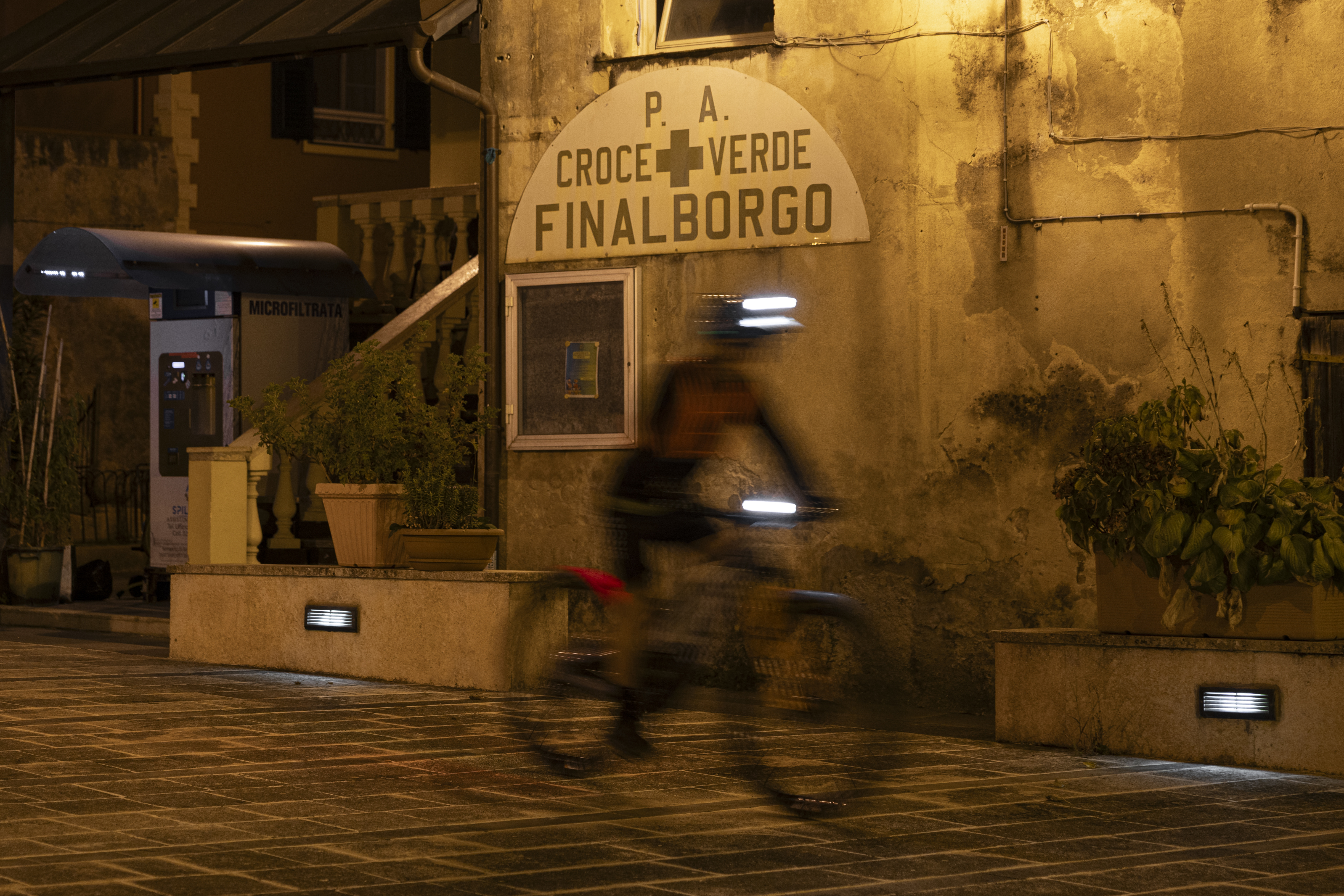 In front of a lit-up building in Finalborgo, Matthew Fairbrother speeds by on his bike with Ergon saddle and grips. The warm light contrasts with the nighttime scene.