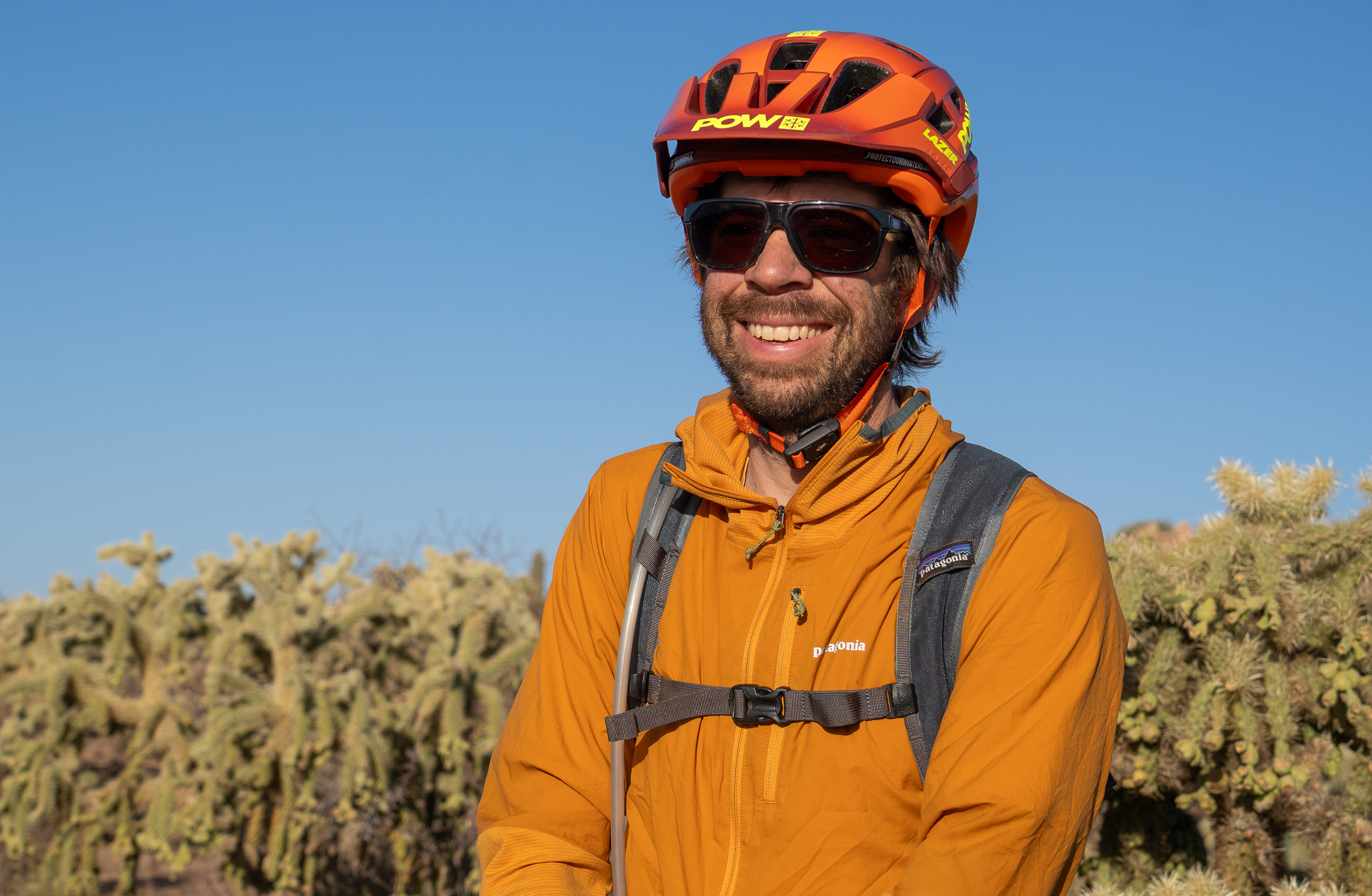 Porträt von Kurt während er lächelt und eine orangene Jacke sowie Fahrradhelm trägt mit Vegetation und Himmel im Hintergrund