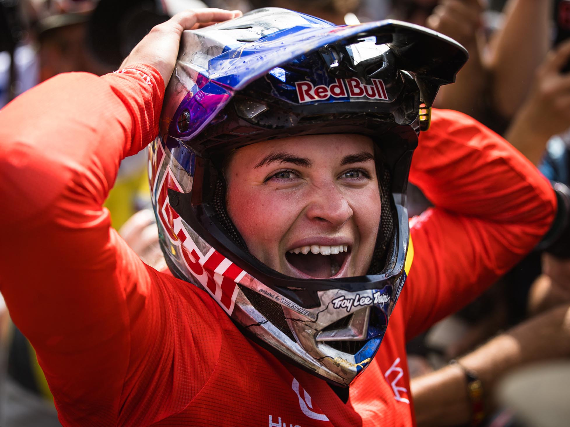 Vali Höll looks excited, wearing a full-face helmet and a red jersey, while spectators cheer in the background.