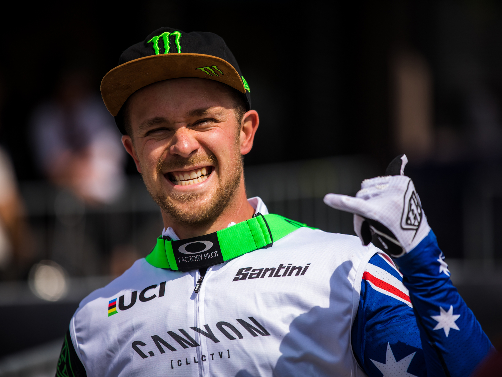 Troy Brosnan lächelt in die Kamera, zeigt mit der Hand das "Hang Loose" Zeichen und trägt dabei eine Schirmmütze, eine neongrüne Goggle um den Hals sowie ein Jersey mit australischer Flagge.