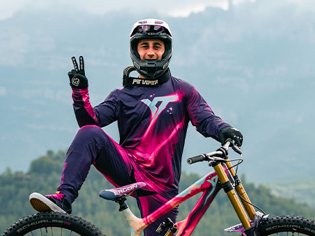 Portrait of Bienvenido Aguado in neon-colored gear and a full-face helmet with his freeride bike. Wooded mountains in the distant background.