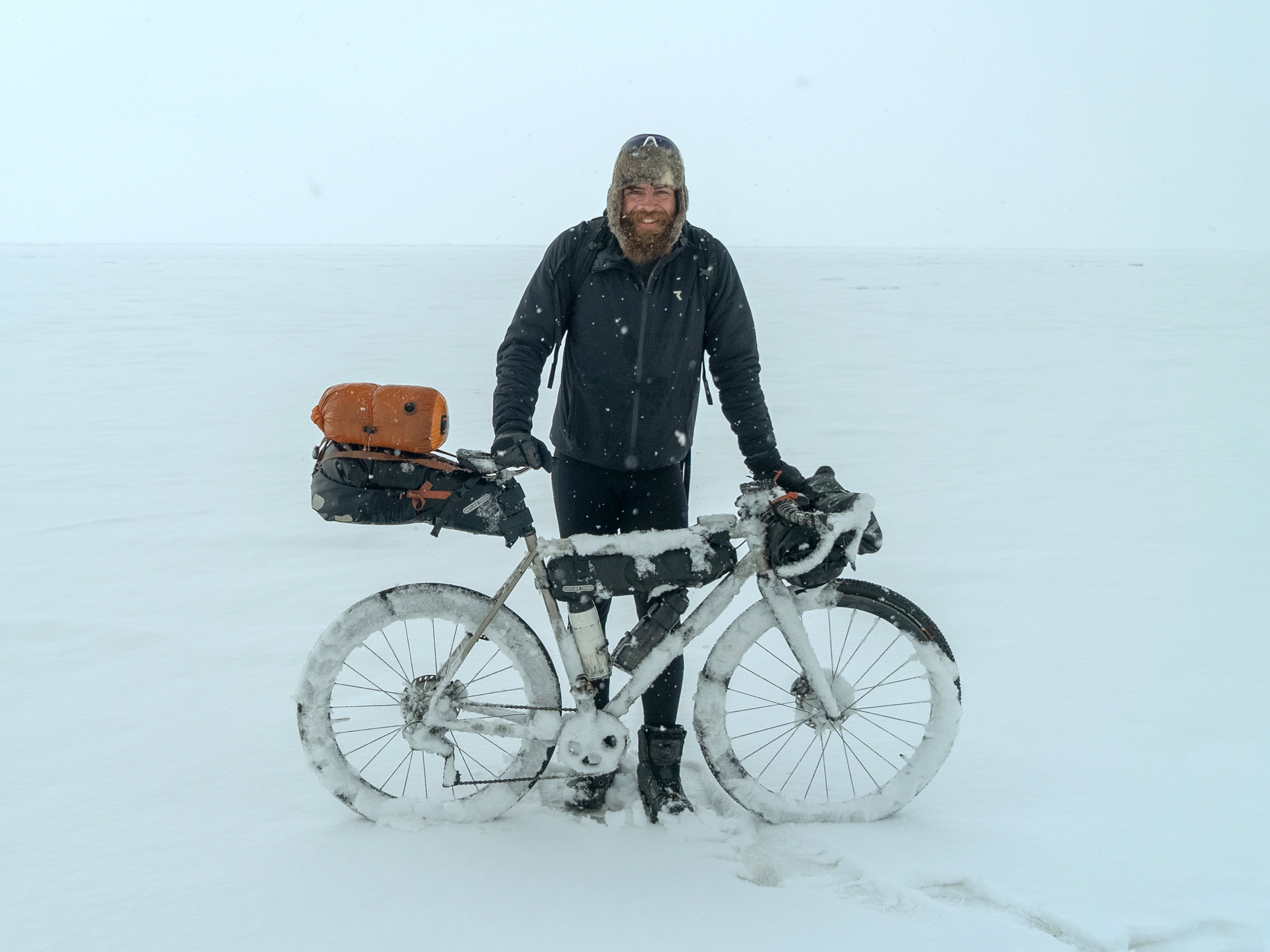 Jonas Deichmann stands smiling with his snow-covered gravel bike, including bike packing equipment, in a snowy landscape, wearing a fur hat.