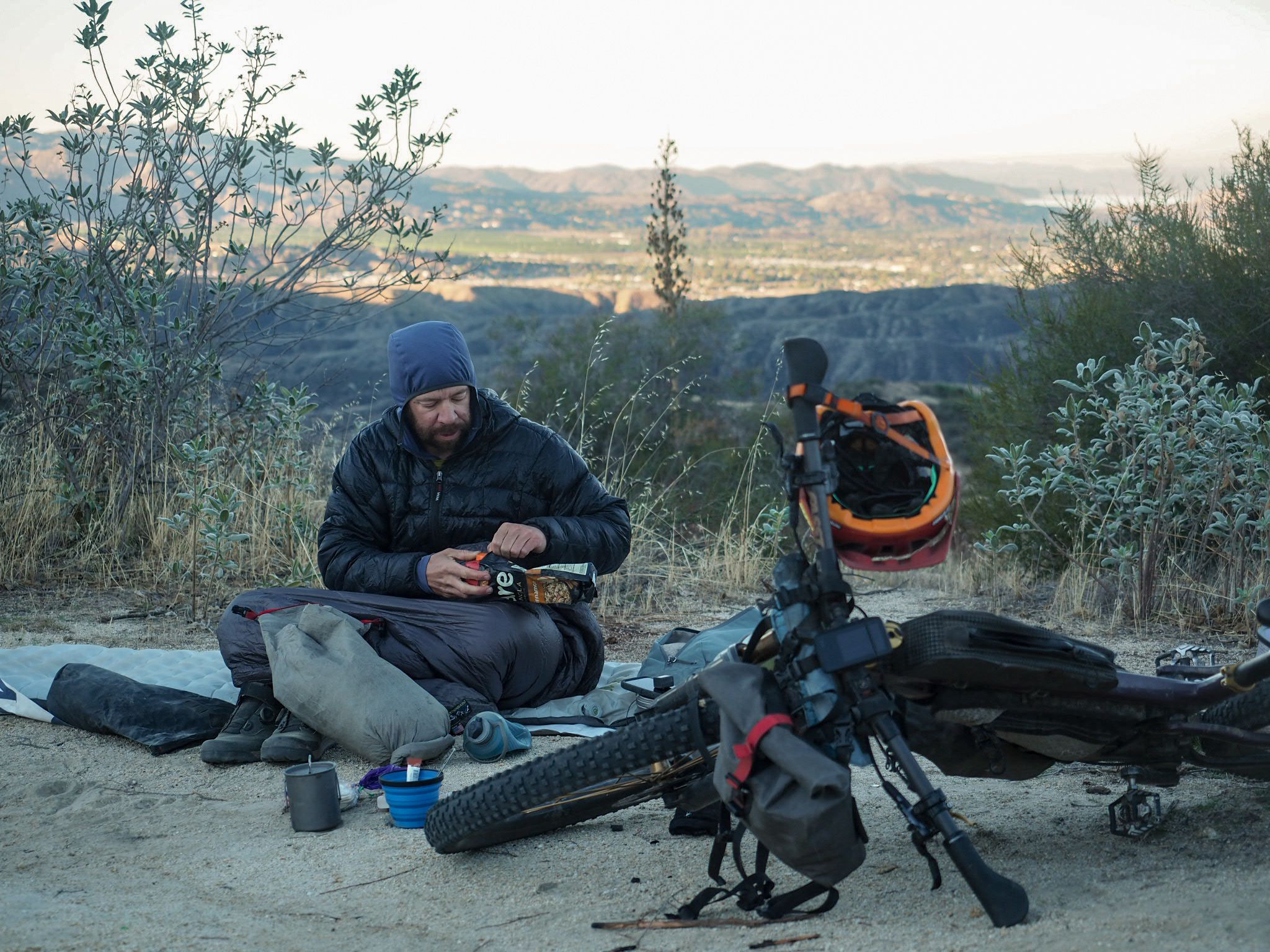 Ein Bikepacker legt eine Pause neben seinem beladenen Fahrrad ein während seines Long-Distance-Bikepacking-Abenteuers in Amerika. Die Szene entlang des Orogenesis Trail zeigt die selbstversorgte Realität mehrtägiger Bikepacking-Touren, geprägt von Ultra-Endurance-Mountainbiking, abgelegenen Trails und weiten Landschaften Nordamerikas.