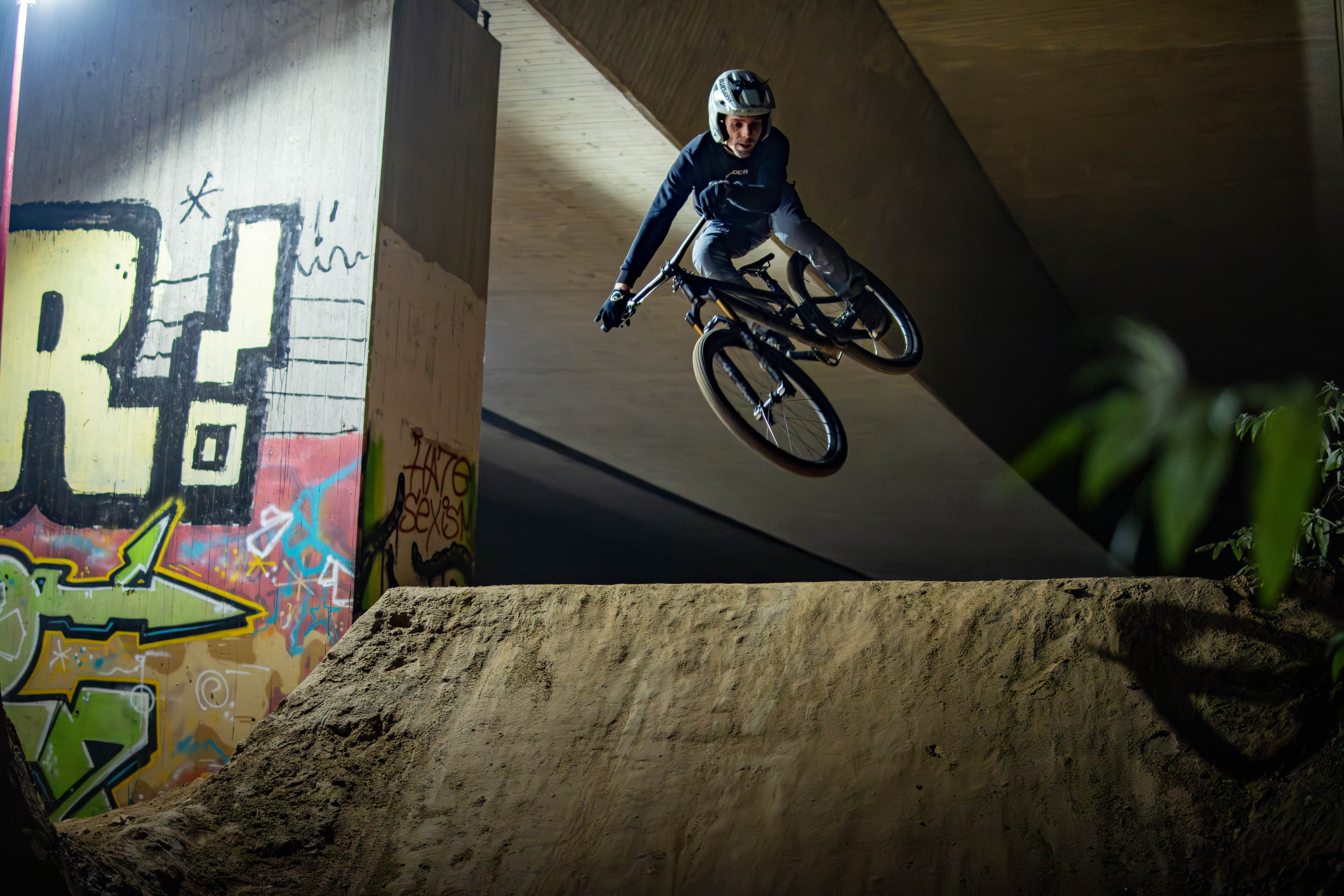 Mountainbike Fahrer bei einem spektakulären Dirt Jump unter einer Brücke. Dynamische Freestyle Action mit urbanem Hintergrund, Sprungtechnik und Bikepark-Atmosphäre.