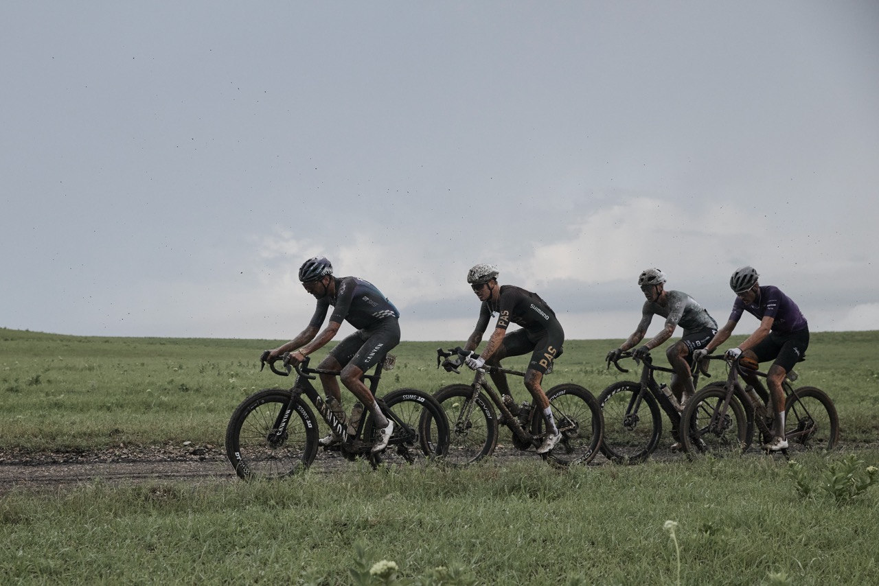 Four gravel racers ride one behind the other on a muddy racecourse