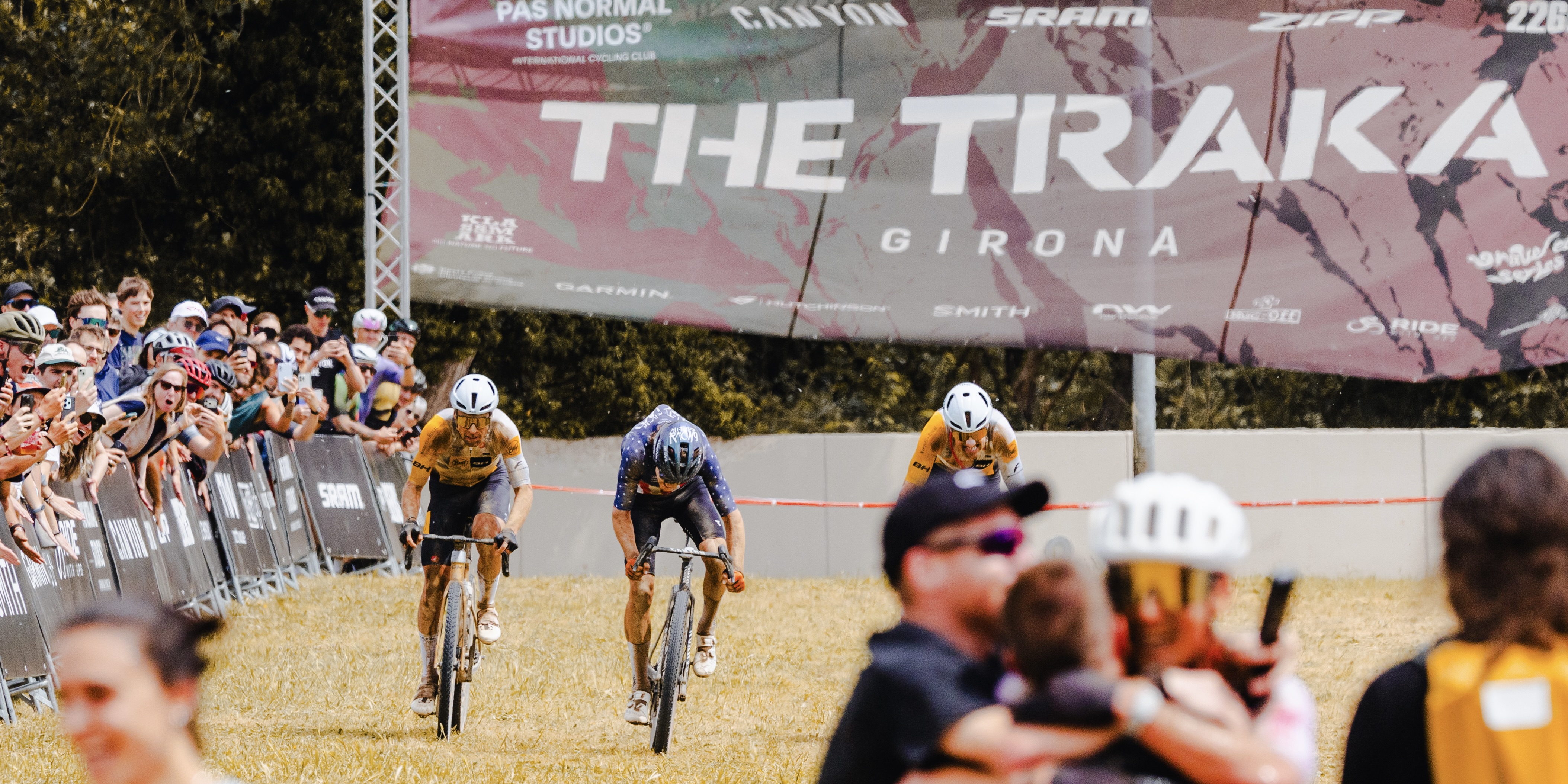 Three participants in the Traka race cross the finish line. Cheering spectators line the course.