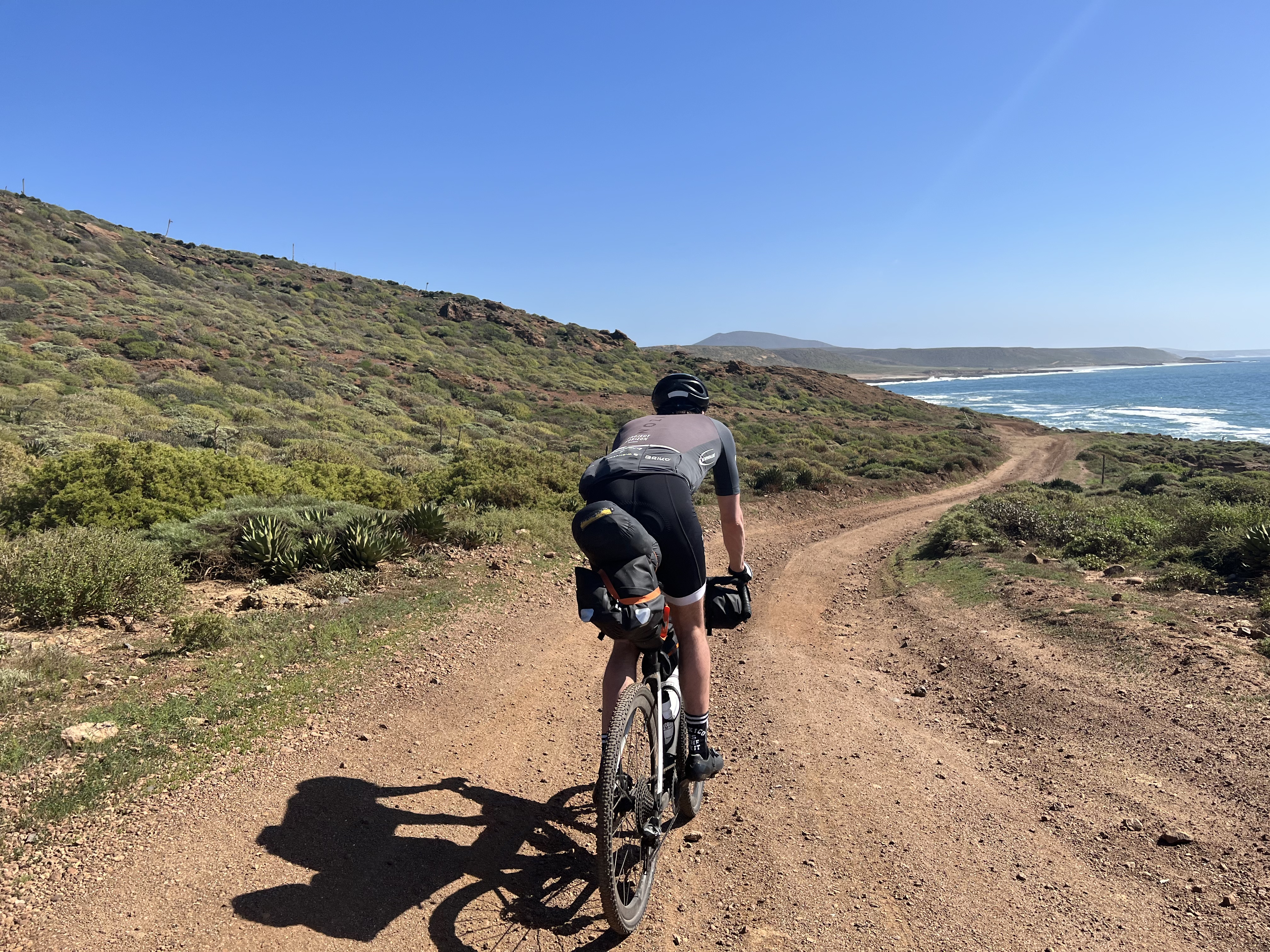 Ein Mann macht eine Bikepacking Tour. Er fährt auf einem Schotterweg. Am Horizont ist das Meer zu sehen.