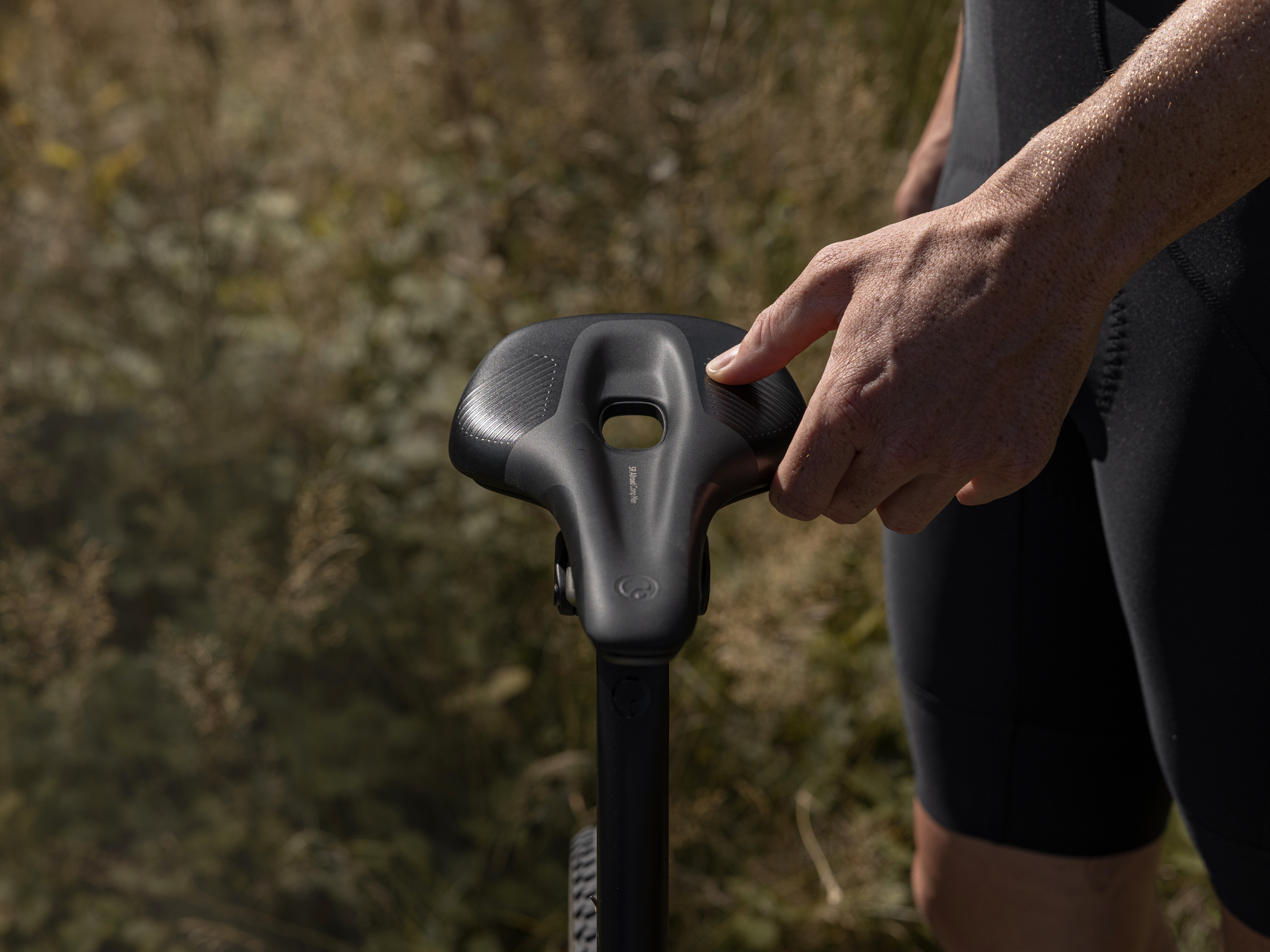 A hand adjusts a bike component near the saddle area in a forest setting