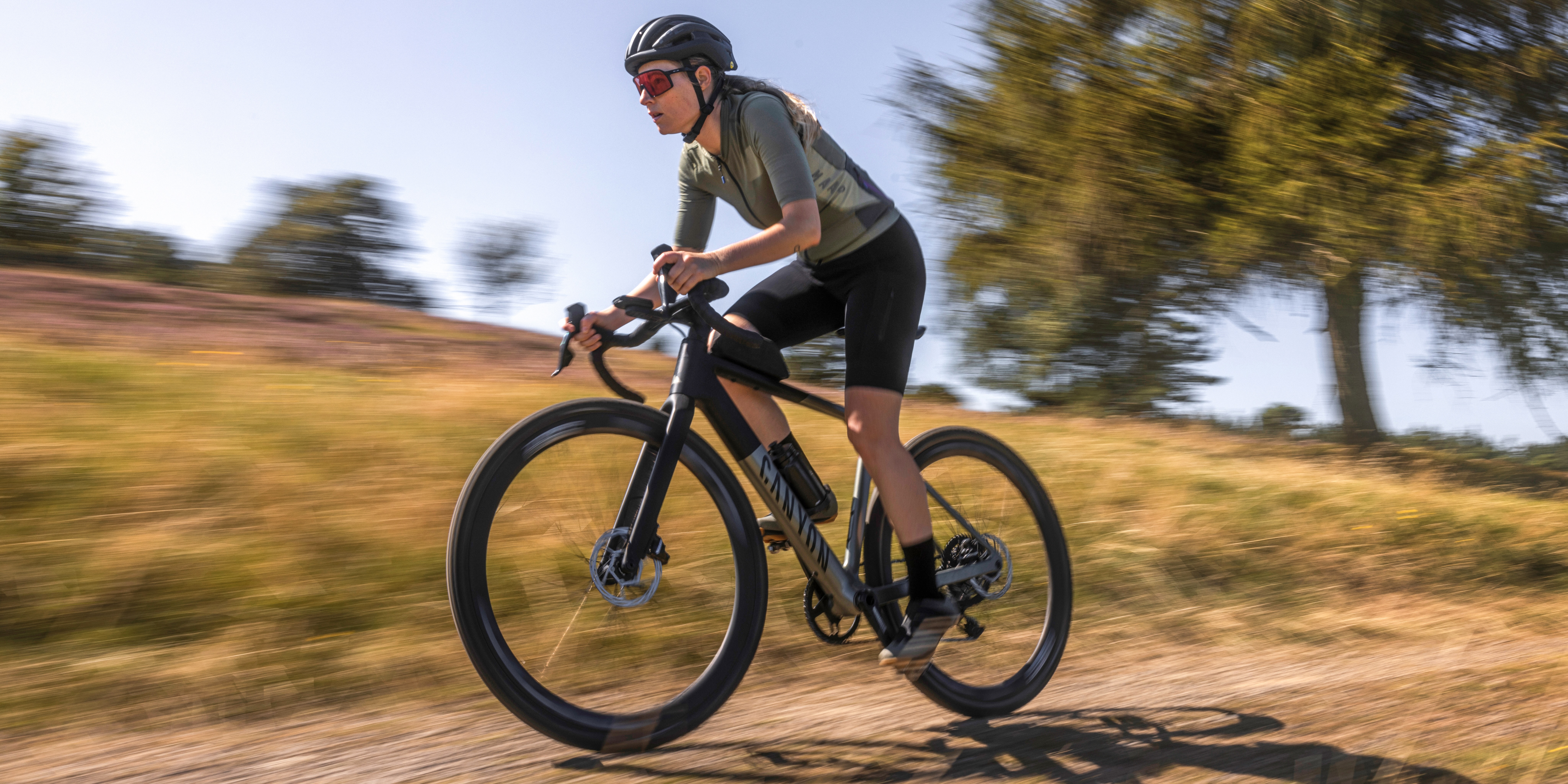 Cyclist rides in a low aerodynamic position on a Canyon gravel bike along a sandy track, background blurred by motion