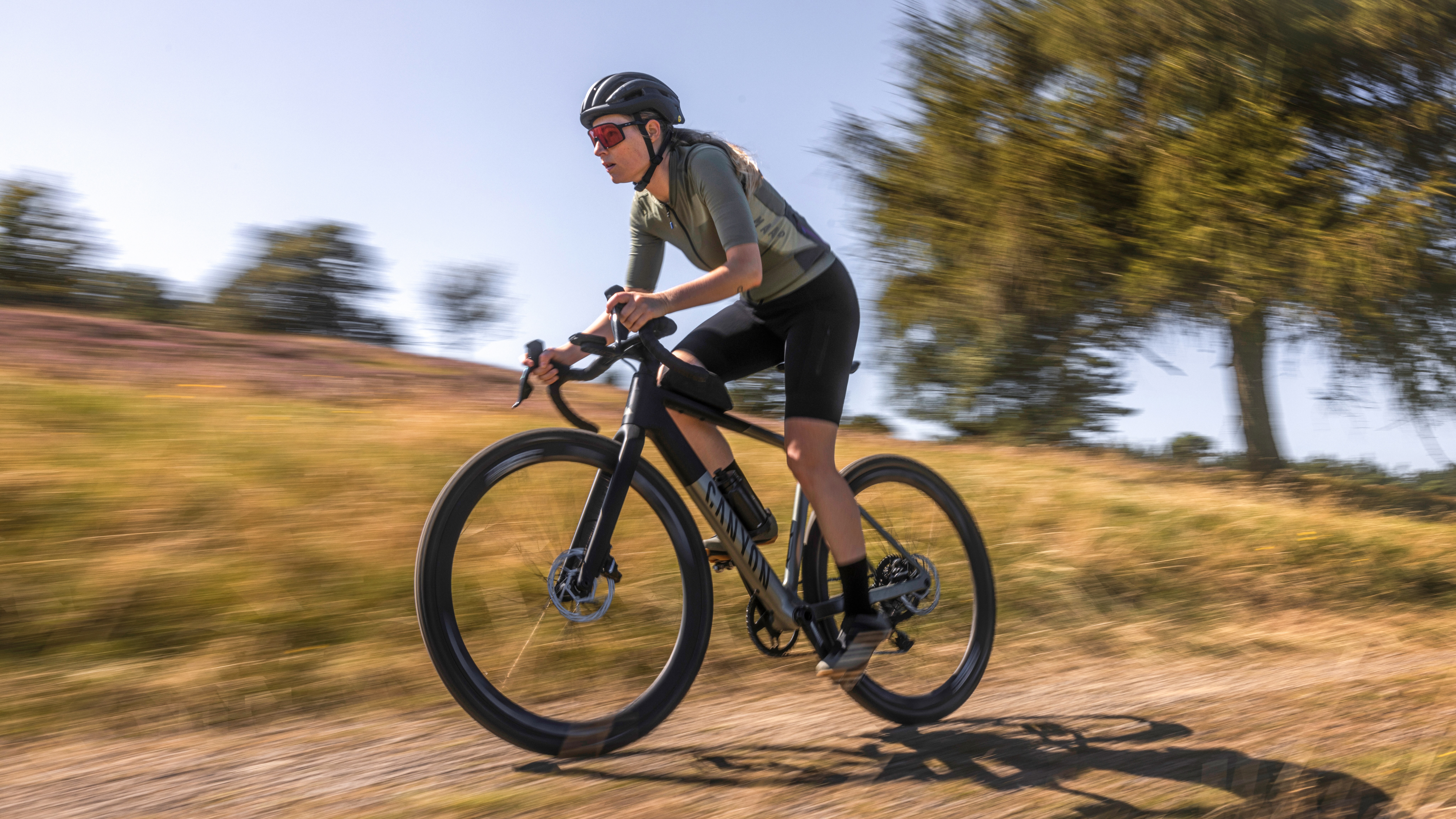 Radfahrerin fährt in geduckter Haltung auf einem Gravelbike über einen sandigen Feldweg, Bewegungsunschärfe im Hintergrund