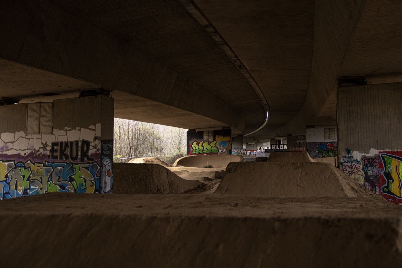 Ein mit Sand aufgeschütteter Pumptrack unter einer Autobahn.