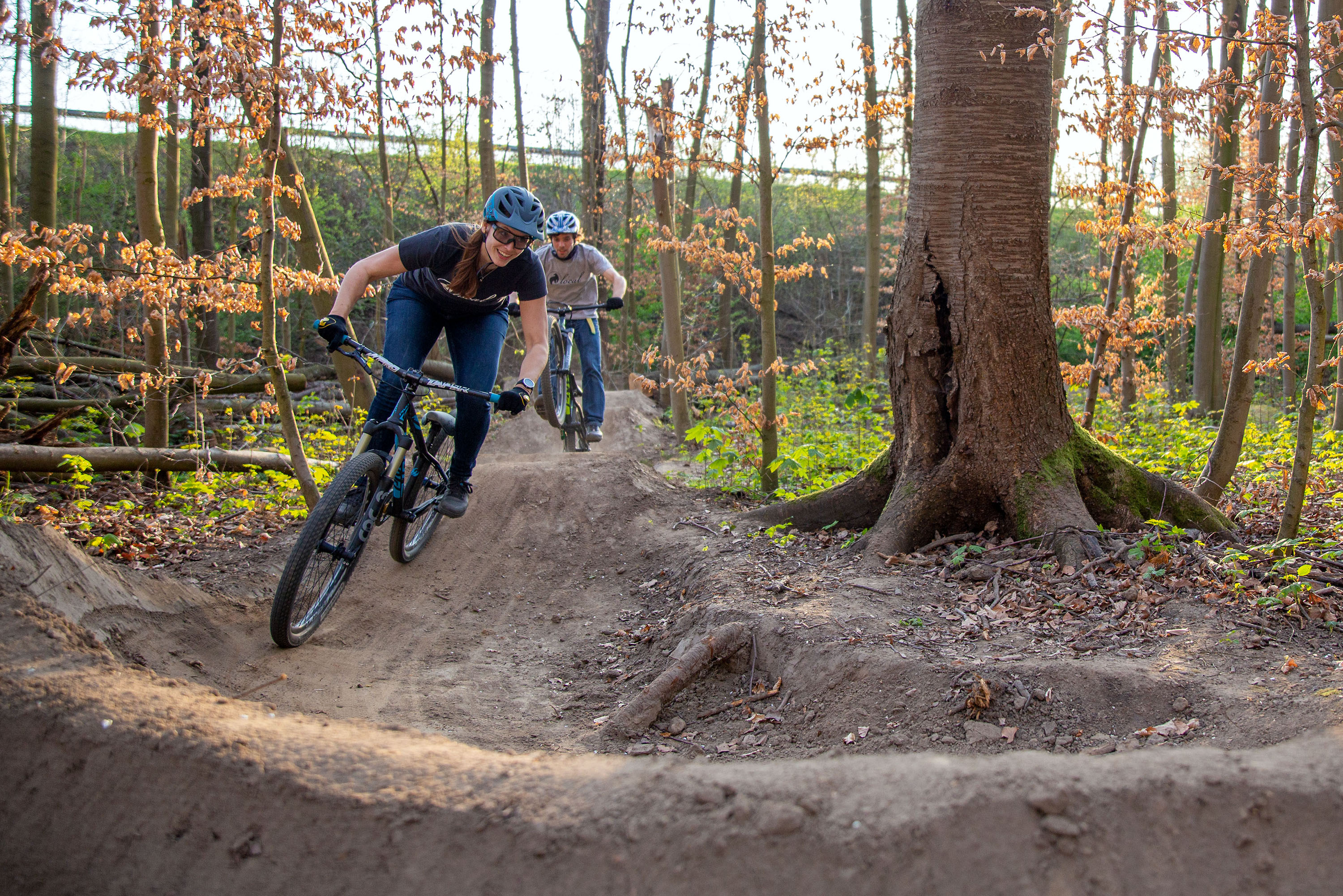 Eine Frau und ein Mann fahren mit MTBs über einen Pumptrack im Wald.