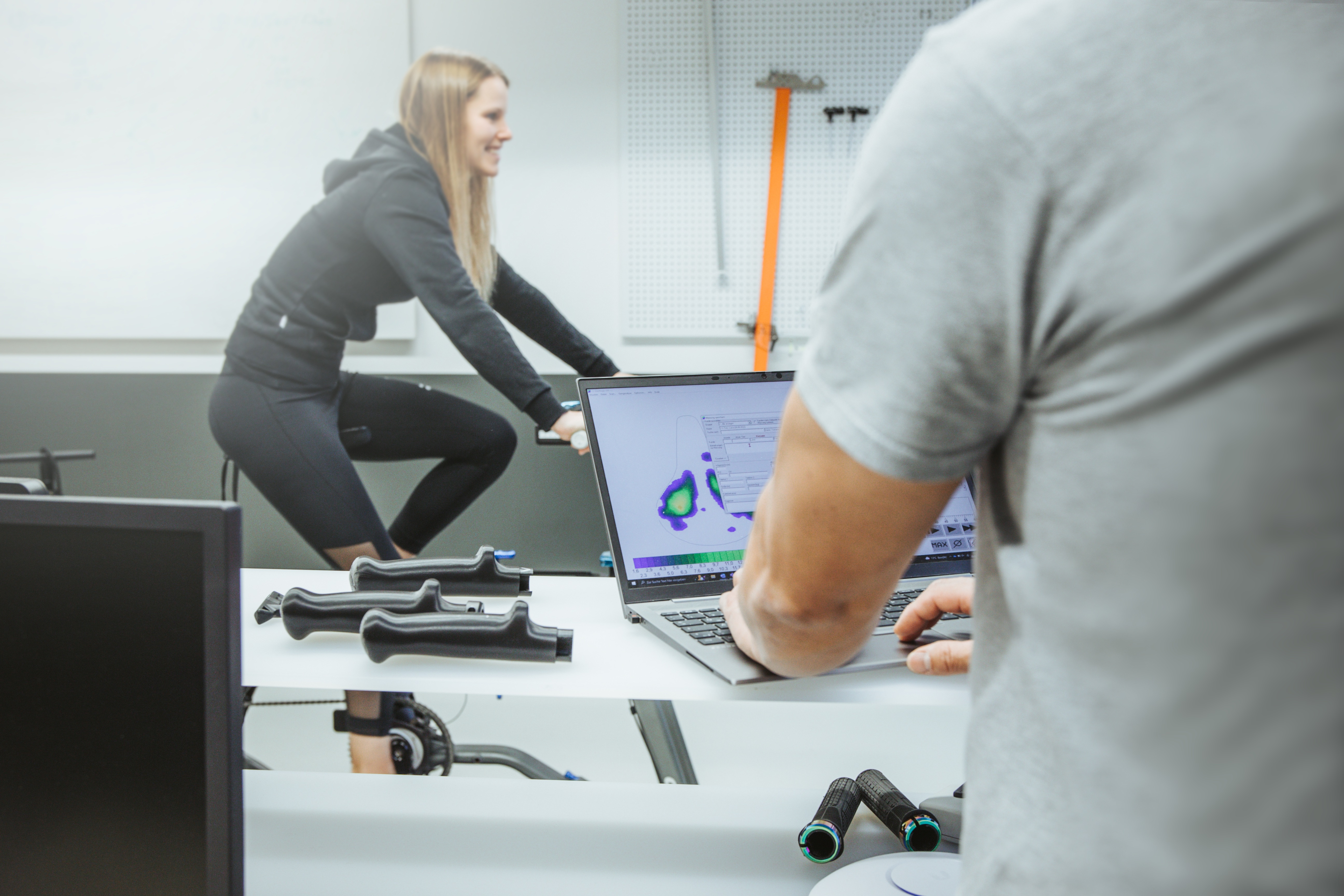 Eine Frau sitzt auf einem Fahrrad während einer Satteldruckmessung. Im Vordergrund wertet Ergonom Kim Tofaute die erfassten Druckdaten am Laptop aus
