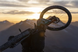 Matthew Fairbrother trägt sein Bike mit Ergon-Komponenten (Sattel und Griffe) auf dem Rücken, während die Sonne über den Berggipfeln aufgeht.